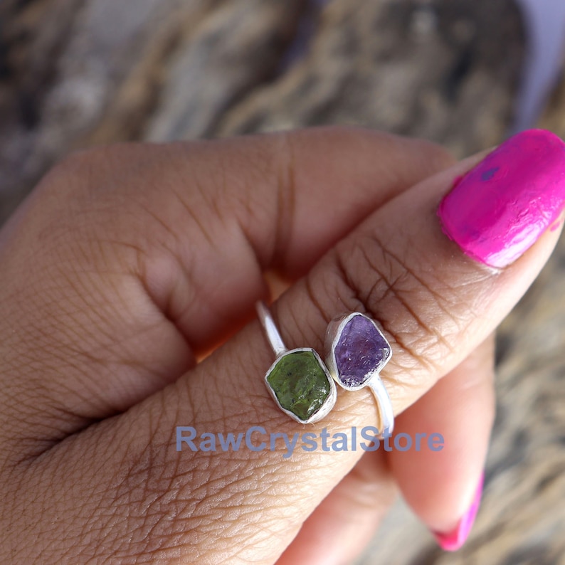 May include: A silver ring with two raw gemstones. The ring features a green gemstone on the left and a purple gemstone on the right. The ring is on a person's finger.