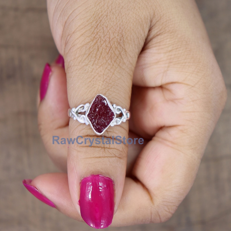 May include: A silver ring featuring a red gemstone set in a diamond-shaped setting. The ring has a delicate, intricate design.