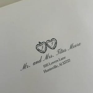 Puede incluir: Un sobre blanco con un sello de tinta negra. El sello presenta dos corazones entrelazados con las iniciales "J" y "T" en su interior. El texto "Mr. and Mrs. Titus Moore 1116 Lovers Lane Huntsville, AL 35757" está impreso debajo de los corazones.