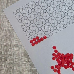 May include: A white paper with a grid of black circles and scattered red, round, gem-like beads. Some beads are placed in the circles, while others are loose. The background is a textured, light gray surface.