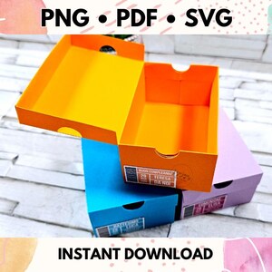 May include: Three colorful boxes with lids, stacked on top of each other. The top box is orange, the middle box is blue, and the bottom box is purple. The boxes have text on them, including "Super Extra Grande" and "Teresa Dani".