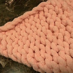 May include: A pink knitted blanket with a fluffy texture. The blanket is made with a chunky yarn and has a unique stitch pattern.