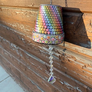 May include: A terracotta flower pot painted with colourful dots and a crystal prism hanging from a chain. The pot is hanging from a wooden wall.