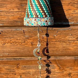 Puede incluir: Un carillón de maceta de terracota con un patrón de lunares marrones, blancos y verde azulado. El carillón tiene una cadena plateada con una luna creciente y cuentas de cristal.