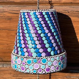Puede incluir: Un carillón colgante hecho de una maceta de terracota pintada. La maceta está decorada con un patrón de puntos azules, morados y blancos. El carillón tiene un cristal colgando de la parte inferior.