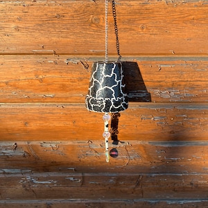 May include: A black and white patterned terracotta pot wind chime hanging from a silver chain. The pot has a white and black geometric pattern and is decorated with iridescent beads.