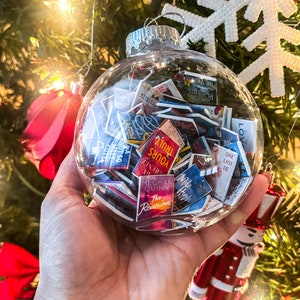 May include: A clear glass Christmas ornament filled with miniature book covers. The ornament is held in a hand, with a Christmas tree and decorations in the background. The book covers have titles like "The Reunion" and "One Last Lie".