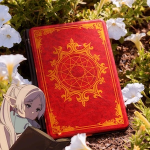 May include: A red book with gold ornate designs, including a central starburst pattern, rests among white flowers and green foliage. The book's cover is a vibrant red, with gold accents. The image evokes a fantasy or magical theme.