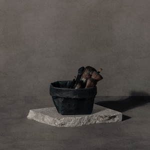 May include: A black paper bag filled with makeup brushes sits on a gray stone platform. The brushes are various sizes and shapes, with black and brown handles.