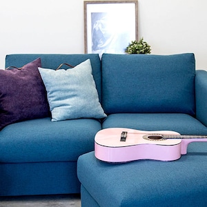 May include: A teal blue sectional sofa with two throw pillows, one purple and one light blue. A pink acoustic guitar is resting on the ottoman in front of the sofa.