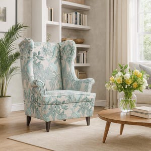 May include: Wingback armchair with a light blue and white tropical leaf pattern. The chair has a high back, wooden legs, and is placed in a living room setting. A round wooden coffee table and a vase of flowers are in front of the chair.