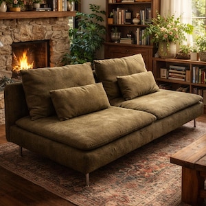 May include: An olive green sofa with several cushions, set in a living room. The sofa has a low design and rests on silver metal legs. The background features a fireplace, bookshelves, and plants, creating a comfortable atmosphere.