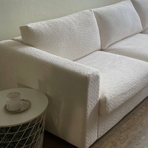 May include: A white plush sofa with a textured fabric. The sofa has two cushions and a thick armrest. The sofa is sitting on a wooden floor.