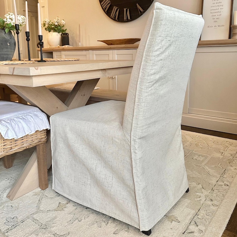 May include: A beige linen dining chair slipcover with a simple design. The slipcover is made of a soft, textured fabric and fits snugly over the chair.