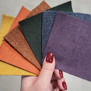Op de afbeelding: Een hand toont een waaier van stoffen stalen in verschillende kleuren. De stalen omvatten tinten geel, oranje, bruin, groen, marineblauw en paars. De stoffen hebben een getextureerd uiterlijk, wat wijst op bekledings- of gordijnmaterialen.