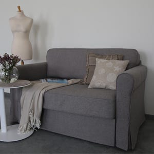 May include: A gray fabric sofa with two pillows, one brown and one beige with a white seashell design. The sofa has a blanket draped over it and is in a room with a white wall and a small white table.