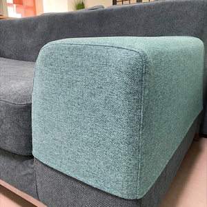May include: A close-up of a teal blue sofa with a dark grey fabric armrest. The sofa has a textured fabric with a subtle pattern.