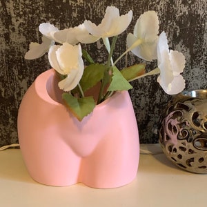 May include: A pink ceramic vase shaped like a human body, with a bouquet of white flowers inside.