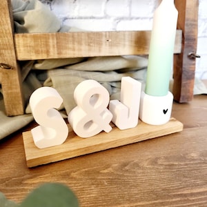 Könnte beinhalten: Weiße Holzbuchstaben, die "S&J" buchstabieren, mit einer grünen Kerze in einem weißen Halter auf einer Holzbasis.