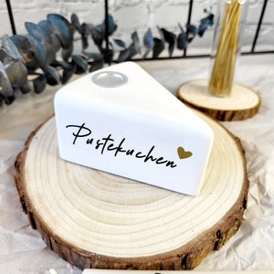 May include: White ceramic cake slice candle holder with the text "Pustekuchen" and a gold heart. The holder is sitting on a wooden slice.