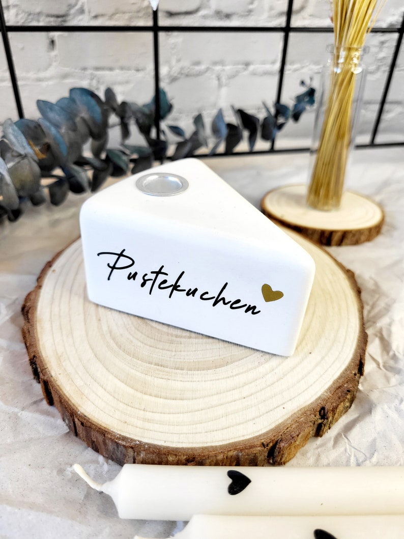 May include: White ceramic cake slice candle holder with the text "Pustekuchen" and a gold heart. The holder is sitting on a wooden slice.