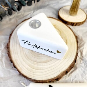 May include: White ceramic cake slice candle holder with a gold heart and the text "Pustekuchen" on a wooden slice. Two white candles with black hearts are in the background.