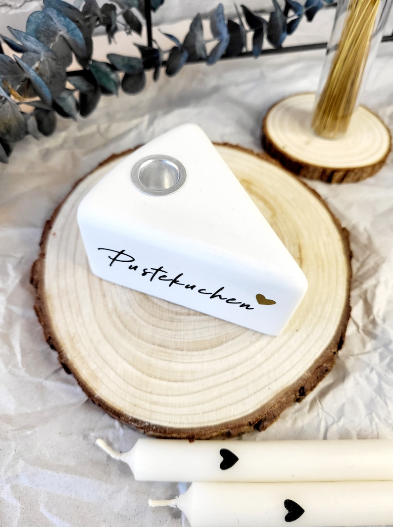May include: White ceramic cake slice candle holder with a gold heart and the text "Pustekuchen" on a wooden slice. Two white candles with black hearts are in the background.