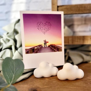 Könnte beinhalten: Eine weiße Fotokarte mit einem Bild von einem Paar, das auf einer Holzbrücke steht. Der Himmel ist leuchtend lila mit einer Herzform, die von einer Vogelschar gebildet wird. Das Paar hält einen roten Ballon. Die Karte wird von zwei weißen wolkenförmigen Haltern gehalten.