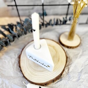 May include: White ceramic cake slice with the text "Pustekuchen" and a black heart, on a wooden base with a white candle. Two additional white candles with black hearts are on a white surface.