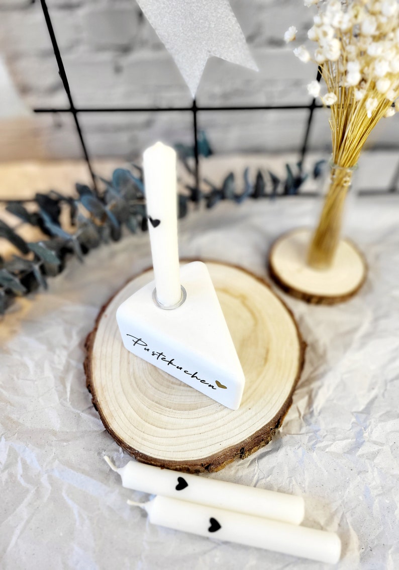 May include: White ceramic cake slice with the text "Pustekuchen" and a black heart, on a wooden base with a white candle. Two additional white candles with black hearts are on a white surface.