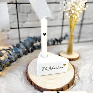May include: White ceramic cake slice candle holder with the word "Pustekuchen" written in gold script. The holder is on a wooden slice and has a white candle with a black heart on it.