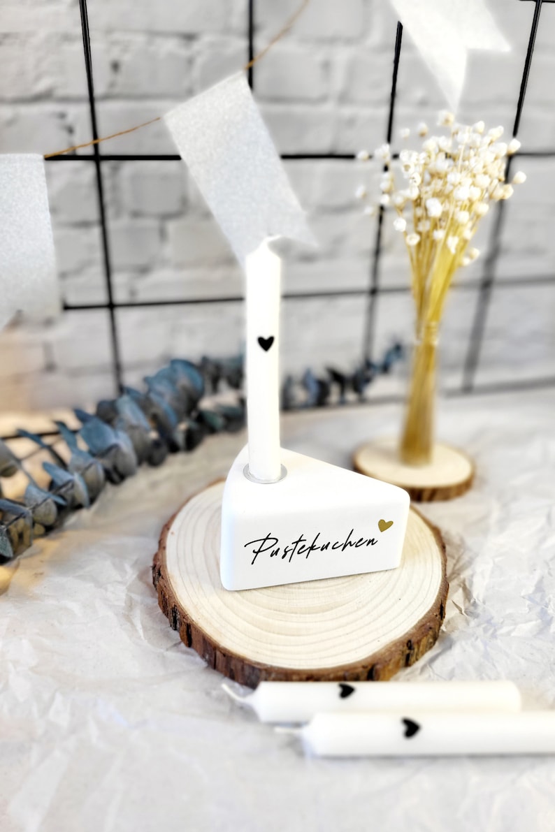 May include: White ceramic cake slice candle holder with the word "Pustekuchen" written in gold script. The holder is on a wooden slice and has a white candle with a black heart on it.