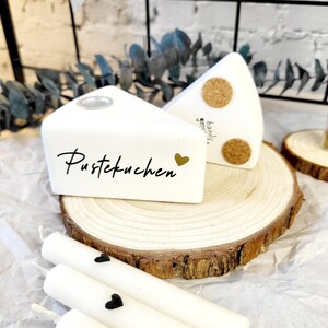 May include: Two white ceramic cake slice candle holders with the words "Pustekuchen" and "handgemacht" on them. The candle holders are sitting on a wooden slice and there are three white candles with black hearts on them.