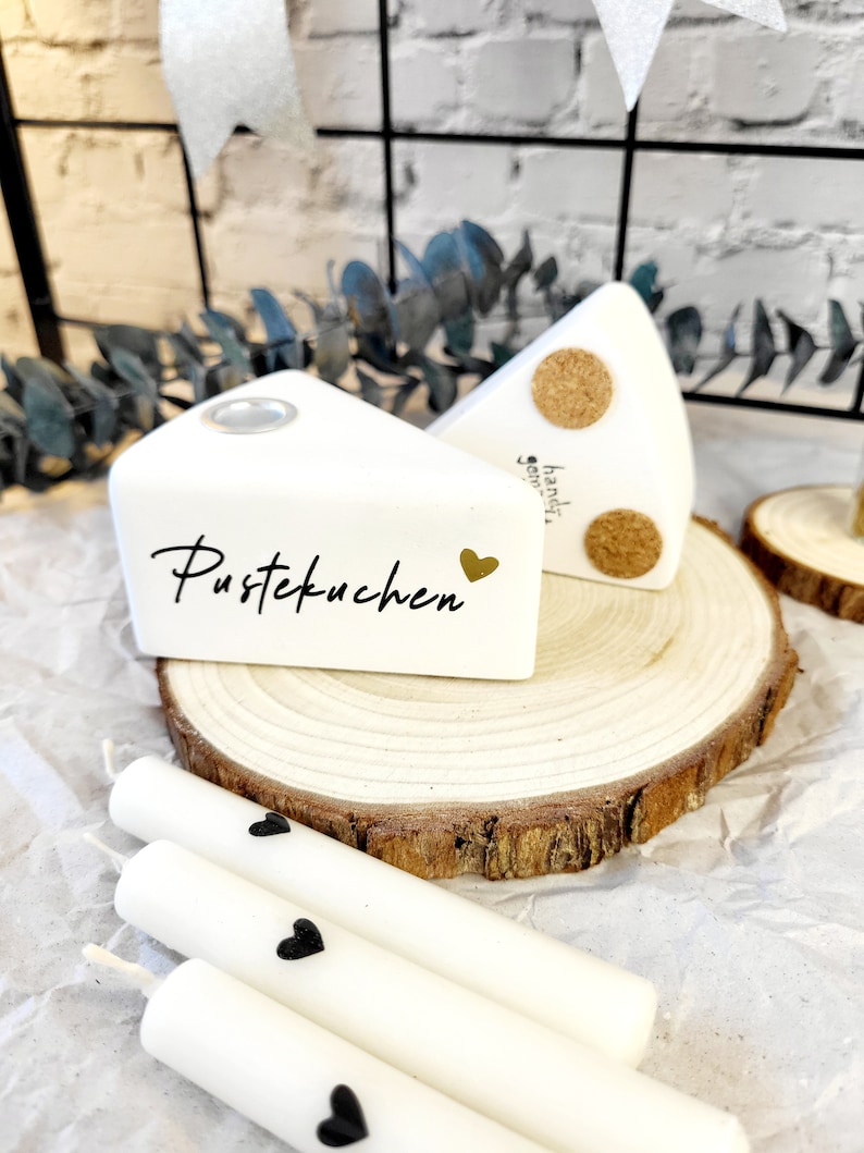 May include: Two white ceramic cake slice candle holders with the words "Pustekuchen" and "handgemacht" on them. The candle holders are sitting on a wooden slice and there are three white candles with black hearts on them.