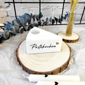 May include: White ceramic cake slice with the text "Pustekuchen" and a gold heart on a wooden slice. Three white candles with black hearts are on a white surface.