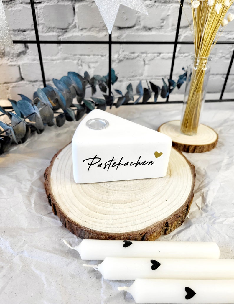 May include: White ceramic cake slice with the text "Pustekuchen" and a gold heart on a wooden slice. Three white candles with black hearts are on a white surface.