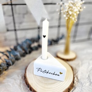 May include: White ceramic cake slice candle holder with the text "Pustekuchen" and a gold heart. The holder is on a wooden slice and a white candle with a black heart is in the holder.