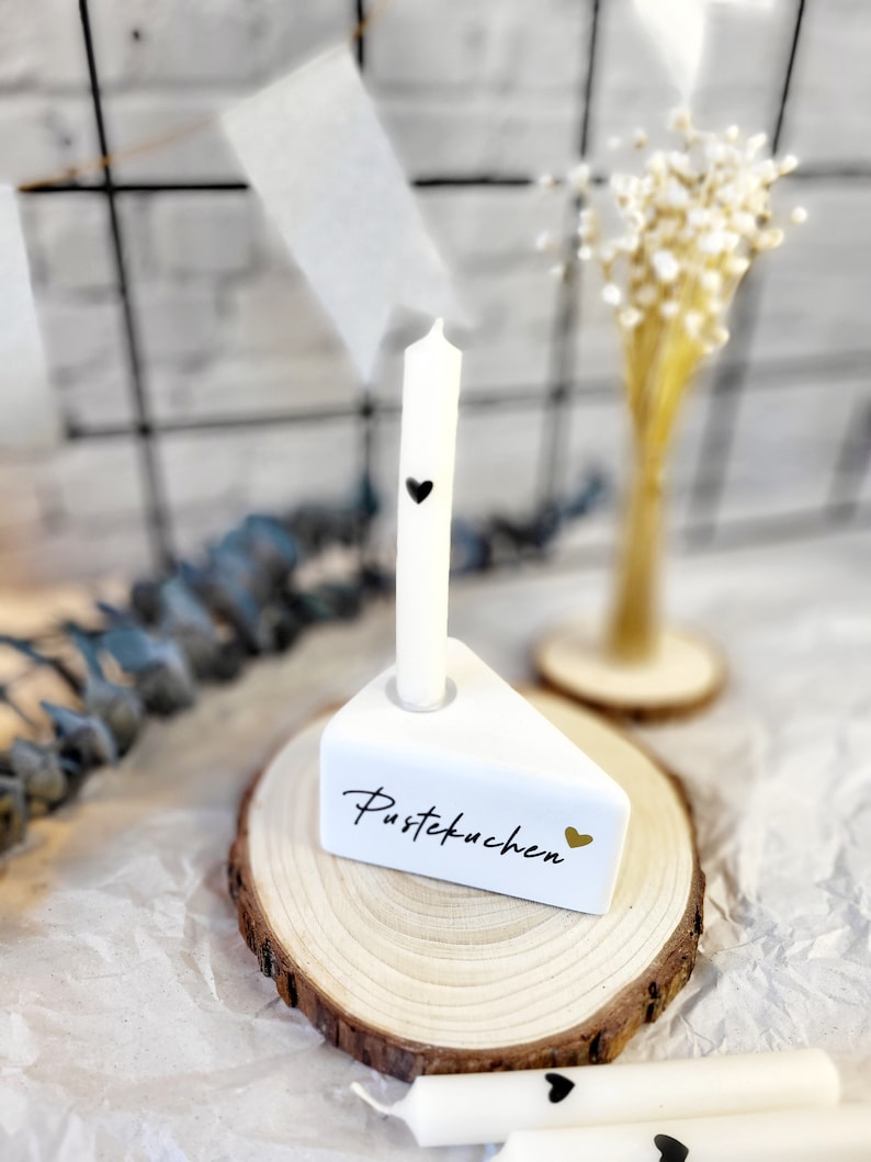 May include: White ceramic cake slice candle holder with the text "Pustekuchen" and a gold heart. The holder is on a wooden slice and a white candle with a black heart is in the holder.