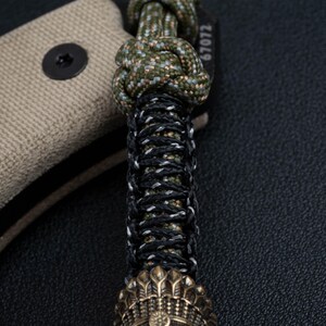 May include: A close-up of a green, black, and white paracord bracelet with a brass bead at the end. The bracelet is tied with a knot and has the number "67072" printed on the side.