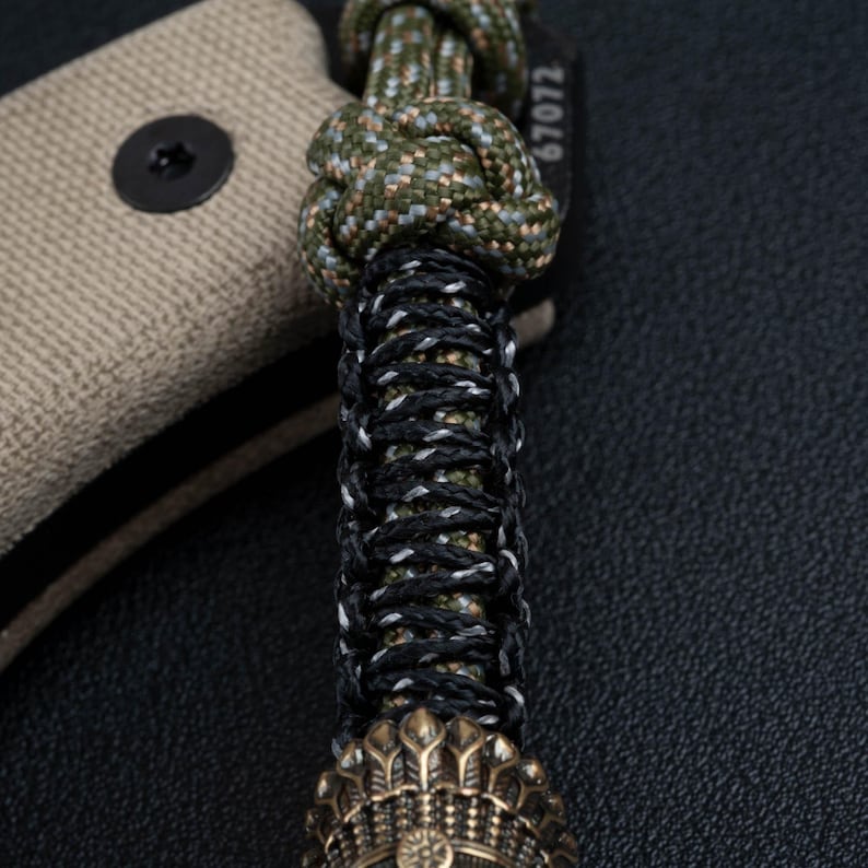 May include: A close-up of a green, black, and white paracord bracelet with a brass bead at the end. The bracelet is tied with a knot and has the number "67072" printed on the side.