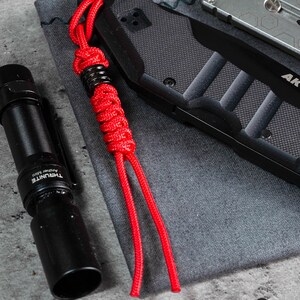 May include: A red paracord bracelet with a black metal bead. The bracelet is lying on a grey fabric surface next to a black torch and a black knife.