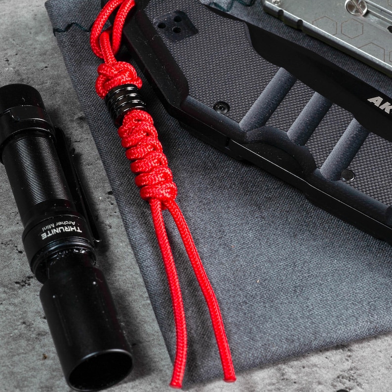 May include: A red paracord bracelet with a black metal bead. The bracelet is lying on a grey fabric surface next to a black torch and a black knife.