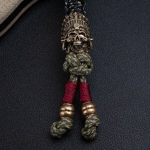 May include: A skull-shaped key chain with a feather headdress, made of brass. The key chain is attached to a green and red paracord.