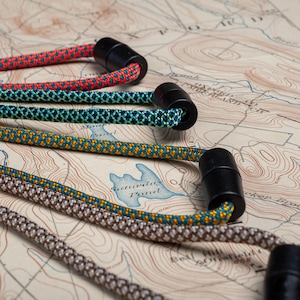 May include: Four different colored paracord lanyards with black plastic clasps. The lanyards are arranged on a map with topographic lines.