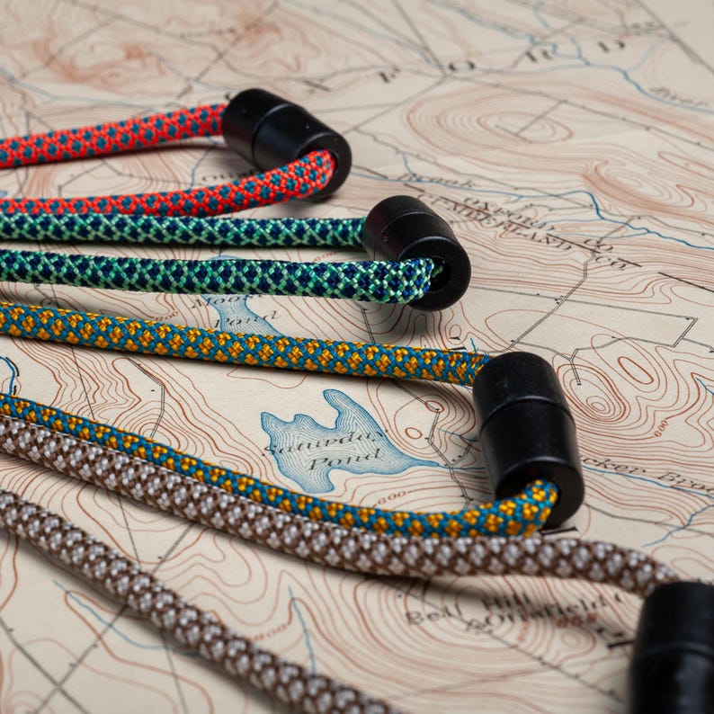 May include: Four different colored paracord lanyards with black plastic clasps. The lanyards are arranged on a map with topographic lines.