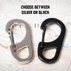 May include: Two metal keychains, one silver and one black, with a hook design. The text "CHOOSE BETWEEN SILVER OR BLACK" is printed above the keychains.