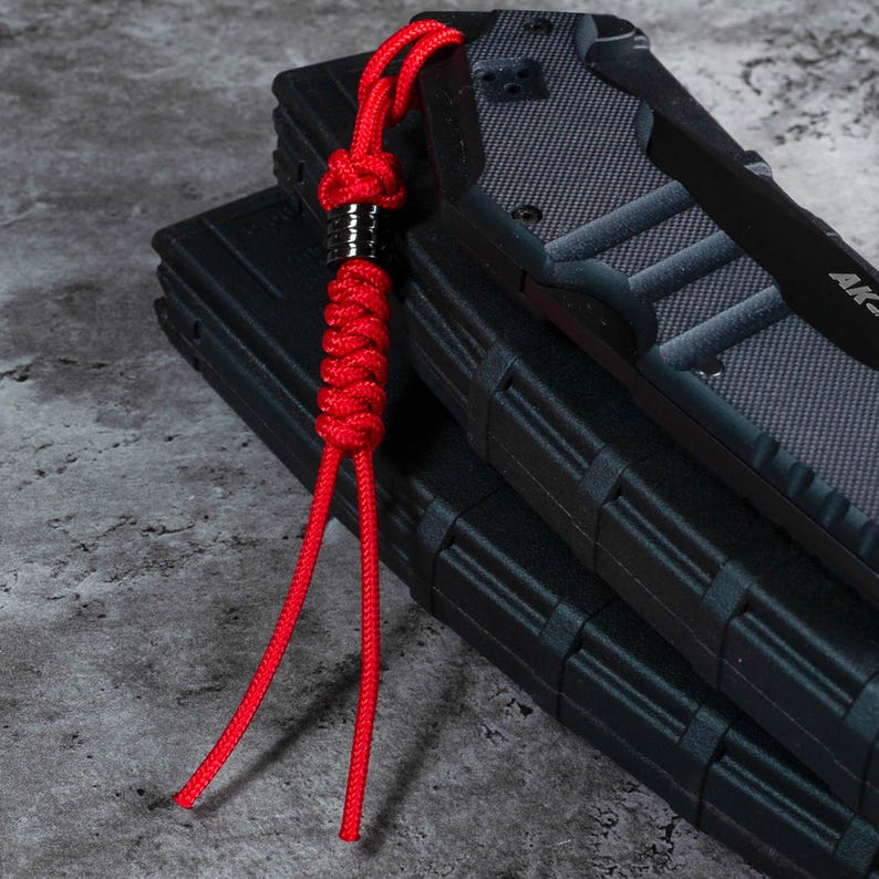 May include: A red paracord lanyard with a black metal bead. The lanyard is attached to a black knife with a textured handle.