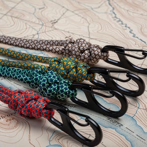 May include: Four colorful paracord keychains with black metal clips. The paracord is knotted and features a variety of patterns, including brown, yellow, green, and red.