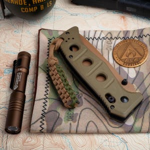 May include: A green and black folding knife with a brown paracord lanyard. The knife is on a camouflage fabric with a copper-colored metal patch that says "Extinction Level Event" and "ELE Survival".