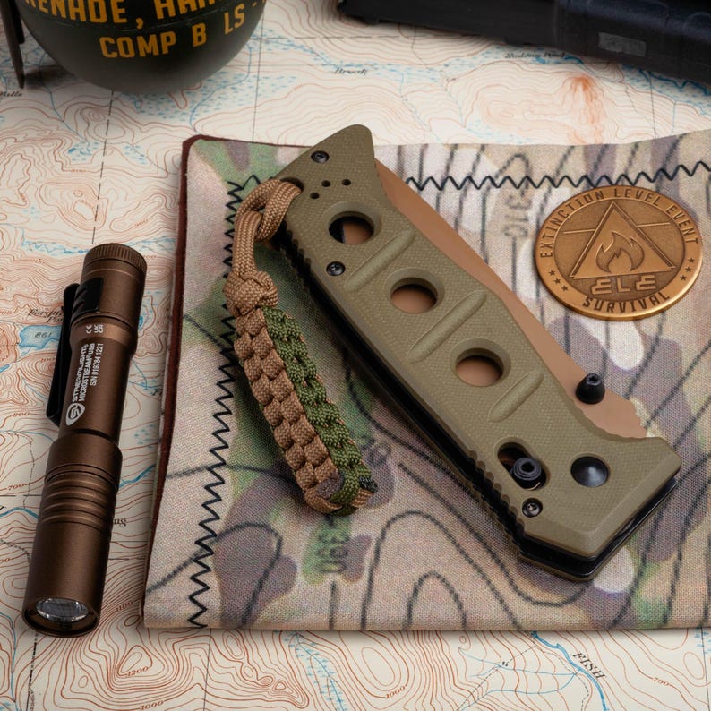 May include: A green and black folding knife with a brown paracord lanyard. The knife is on a camouflage fabric with a copper-colored metal patch that says "Extinction Level Event" and "ELE Survival".